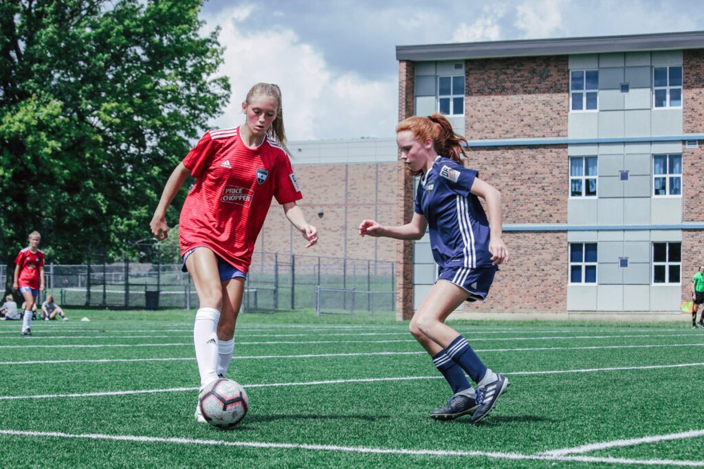 Two teenage girls compete during a soccer match on an outdoor turf field. The player on the left, wearing a red jersey with white socks, controls the ball with her foot while being closely defended by a player in a navy blue uniform. Both girls appear focused and engaged in the game. In the background, a brick and grey school building, a referee, and additional players and spectators are visible under a partly cloudy sky.