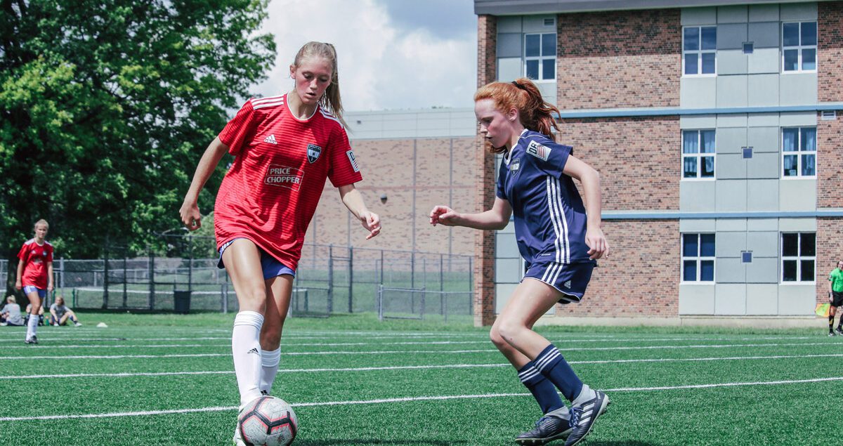 Two teenage girls compete during a soccer match on an outdoor turf field. The player on the left, wearing a red jersey with white socks, controls the ball with her foot while being closely defended by a player in a navy blue uniform. Both girls appear focused and engaged in the game. In the background, a brick and grey school building, a referee, and additional players and spectators are visible under a partly cloudy sky.
