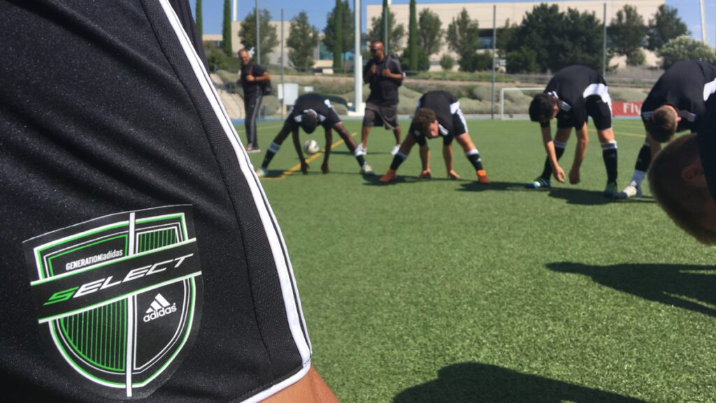 A group of soccer players wearing matching black Adidas uniforms with white stripes are stretching on a green artificial turf field. In the foreground, a close-up of a player’s sleeve prominently displays a patch reading 'GENERATION Adidas SELECT' above the Adidas logo on a black and green shield-shaped emblem. In the background, players are bending forward in a line, touching the ground, with two coaches or trainers observing behind them. The setting appears to be a sunny day at a professional training facility with buildings, trees, and sports equipment in the distance.