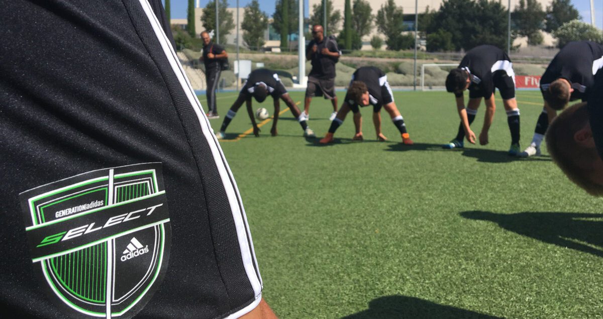 A group of soccer players wearing matching black Adidas uniforms with white stripes are stretching on a green artificial turf field. In the foreground, a close-up of a player’s sleeve prominently displays a patch reading 'GENERATION Adidas SELECT' above the Adidas logo on a black and green shield-shaped emblem. In the background, players are bending forward in a line, touching the ground, with two coaches or trainers observing behind them. The setting appears to be a sunny day at a professional training facility with buildings, trees, and sports equipment in the distance.