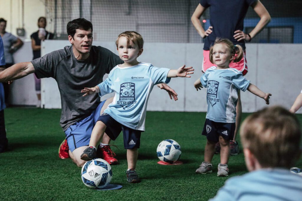A young boy and girl wearing light blue "Sporting Stripes" soccer jerseys and navy shorts stand on an indoor turf field, each with a soccer ball at their feet. The boy has one foot raised and arms outstretched for balance, while a male coach kneels behind him, guiding and encouraging him. The girl, standing nearby, also holds her arms out as if mimicking the pose. Other children and adults are visible in the background, suggesting a youth soccer training session. The atmosphere is playful and instructional.
