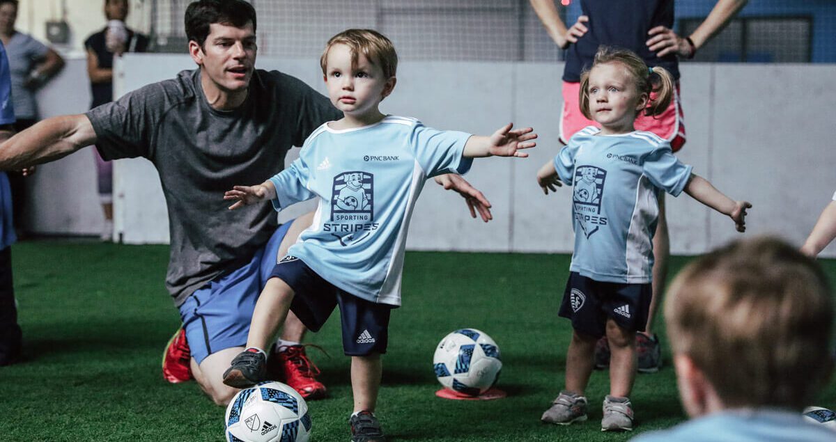 A young boy and girl wearing light blue "Sporting Stripes" soccer jerseys and navy shorts stand on an indoor turf field, each with a soccer ball at their feet. The boy has one foot raised and arms outstretched for balance, while a male coach kneels behind him, guiding and encouraging him. The girl, standing nearby, also holds her arms out as if mimicking the pose. Other children and adults are visible in the background, suggesting a youth soccer training session. The atmosphere is playful and instructional.