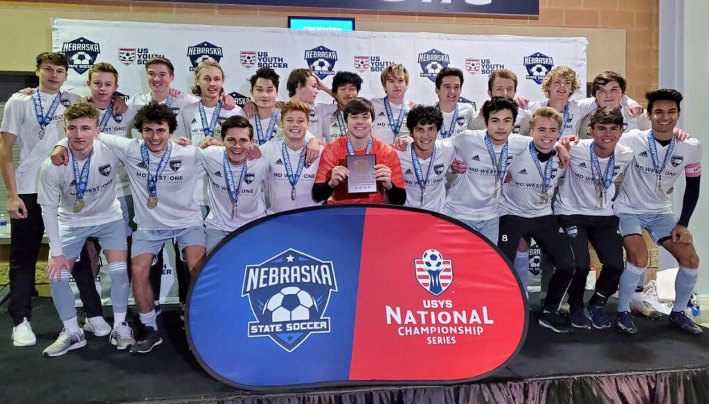 A boys' youth soccer team poses for a group photo on a stage after a tournament victory. They are wearing white jerseys with "MD WEST ONE" printed on the front and silver medals around their necks. One player in a red goalkeeper jersey kneels at the center holding a plaque. Behind them is a backdrop with logos for "Nebraska State Soccer" and "US Youth Soccer." In front of the group is a podium-style sign featuring the Nebraska State Soccer and USYS National Championship Series logos. The players appear joyful and proud, celebrating their achievement together.