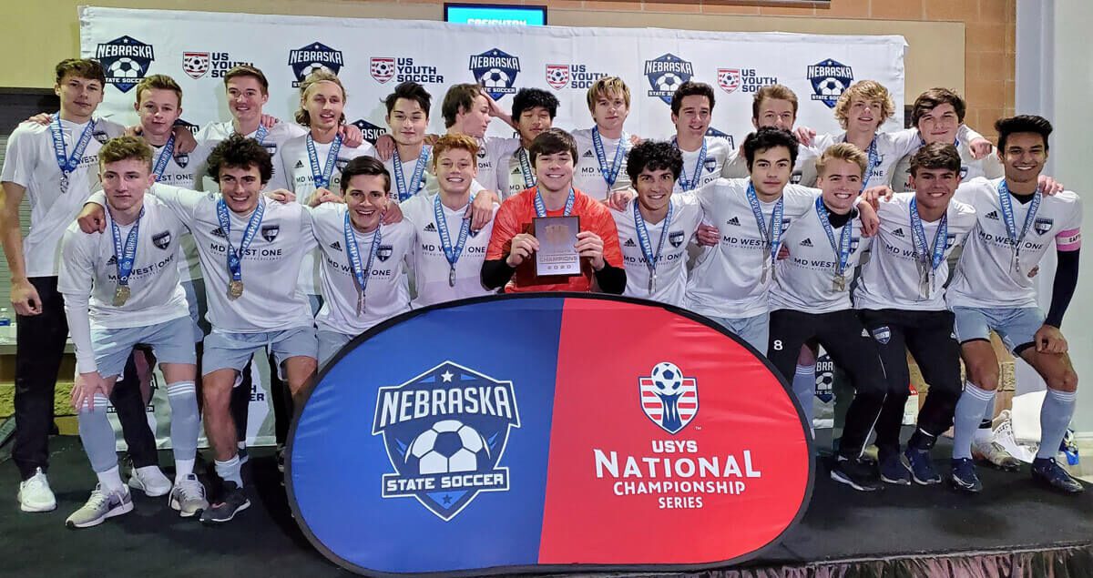 A boys' youth soccer team poses for a group photo on a stage after a tournament victory. They are wearing white jerseys with "MD WEST ONE" printed on the front and silver medals around their necks. One player in a red goalkeeper jersey kneels at the center holding a plaque. Behind them is a backdrop with logos for "Nebraska State Soccer" and "US Youth Soccer." In front of the group is a podium-style sign featuring the Nebraska State Soccer and USYS National Championship Series logos. The players appear joyful and proud, celebrating their achievement together.
