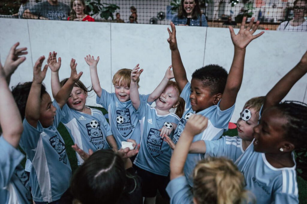 A joyful group of young children wearing matching light blue 'Sporting Stripes' soccer jerseys huddle together indoors, raising their arms high in celebration. They are smiling, laughing, and full of excitement, some with their eyes closed in glee. A coach or adult is partially visible in the center holding something small, possibly a reward or treat. Behind the kids, several adults and parents watch through a low barrier, smiling and enjoying the moment. The setting is an indoor soccer facility with a turf field and netting overhead.
