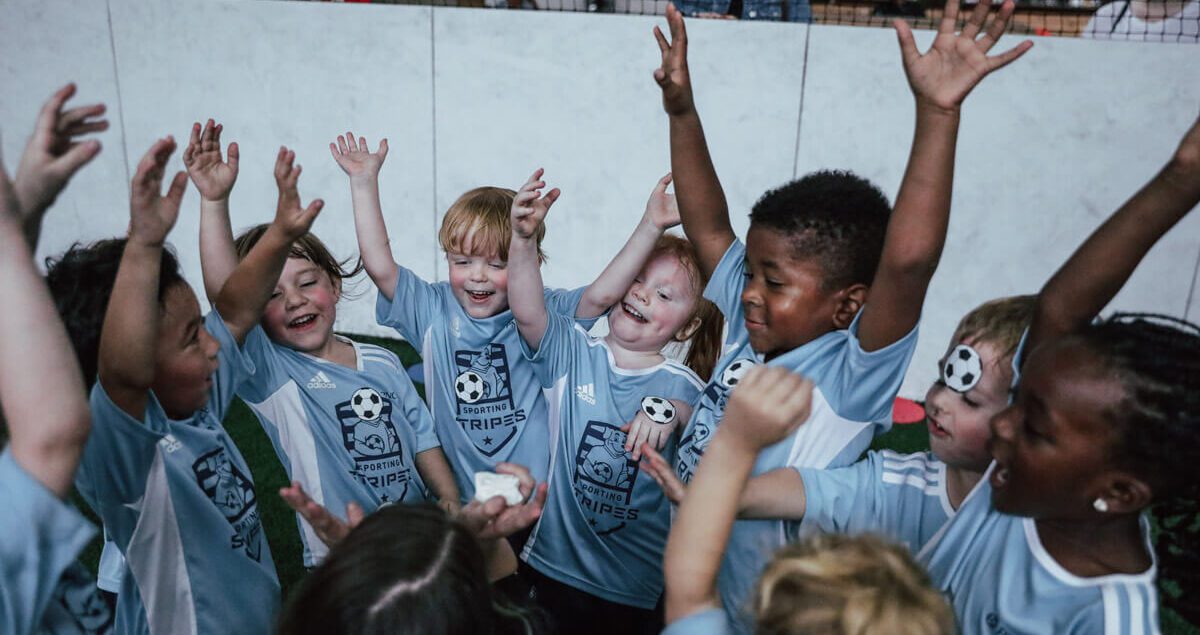 A joyful group of young children wearing matching light blue 'Sporting Stripes' soccer jerseys huddle together indoors, raising their arms high in celebration. They are smiling, laughing, and full of excitement, some with their eyes closed in glee. A coach or adult is partially visible in the center holding something small, possibly a reward or treat. Behind the kids, several adults and parents watch through a low barrier, smiling and enjoying the moment. The setting is an indoor soccer facility with a turf field and netting overhead.