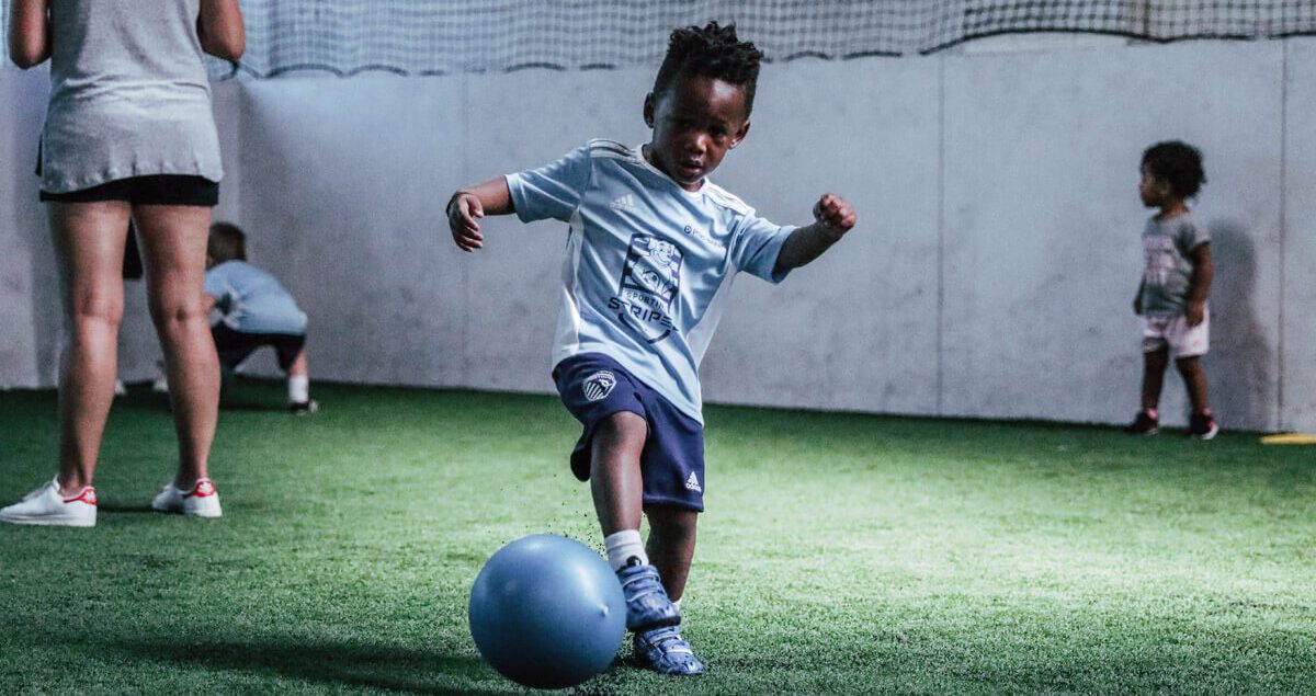 A young child wearing a light blue soccer jersey and navy blue shorts is playing with a blue ball on an indoor turf field. The child is in mid-motion, appearing to kick the ball with focus and determination. In the background, other small children and an adult are also present on the field. The walls are white with a netted upper section, and the lighting highlights the central child’s activity.