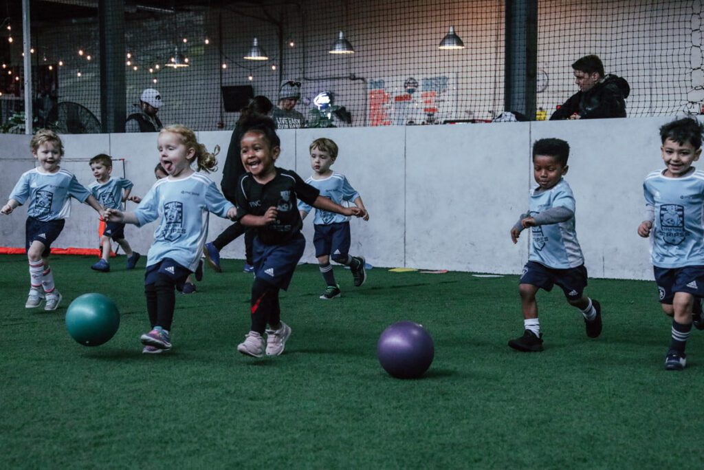 A group of young children, around preschool age, energetically run and play on an indoor turf soccer field. Most of the kids are wearing light blue soccer jerseys with dark blue shorts and athletic shoes, while one girl in the center wears a black jersey. Two colorful balls, one green and one purple, are on the field in front of them. The children are smiling, laughing, and full of energy as they chase the balls. In the background, adults and staff observe from behind a short wall, with netting and string lights adding a warm ambiance to the indoor facility.