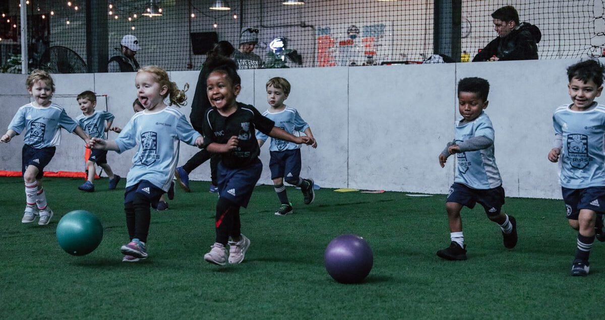 A group of young children, around preschool age, energetically run and play on an indoor turf soccer field. Most of the kids are wearing light blue soccer jerseys with dark blue shorts and athletic shoes, while one girl in the center wears a black jersey. Two colorful balls, one green and one purple, are on the field in front of them. The children are smiling, laughing, and full of energy as they chase the balls. In the background, adults and staff observe from behind a short wall, with netting and string lights adding a warm ambiance to the indoor facility.