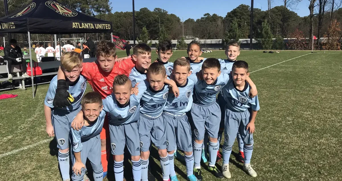 A youth soccer team in matching light blue uniforms poses together on a grassy field, smiling with arms around each other, with a tournament tent and trees in the background.