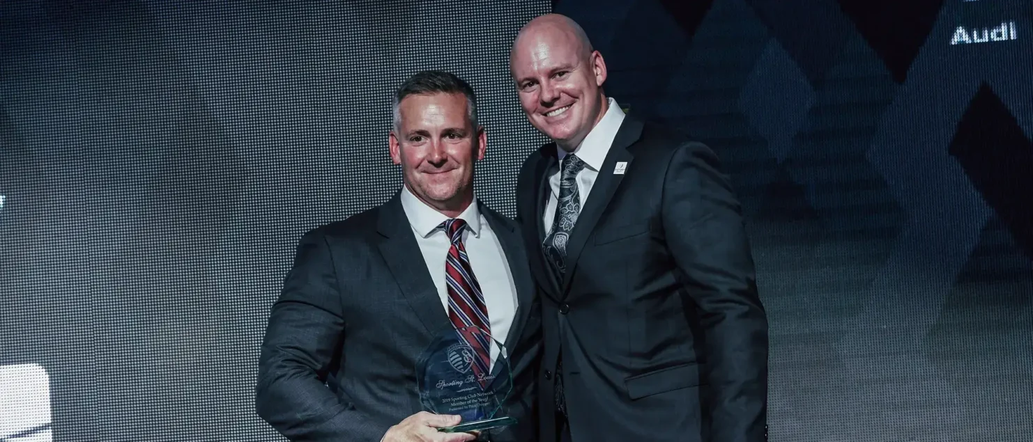 Two men dressed in formal suits stand side by side on a stage in front of a dark, patterned backdrop with corporate branding, including the Audi logo. The man on the left holds a glass award with both hands and smiles confidently, while the man on the right, also smiling, rests his arm around the other’s back. Part of the text on the backdrop reads 'OF THE YEAR,' suggesting this is an award ceremony. The lighting emphasizes their faces and the award, creating a professional and celebratory atmosphere.