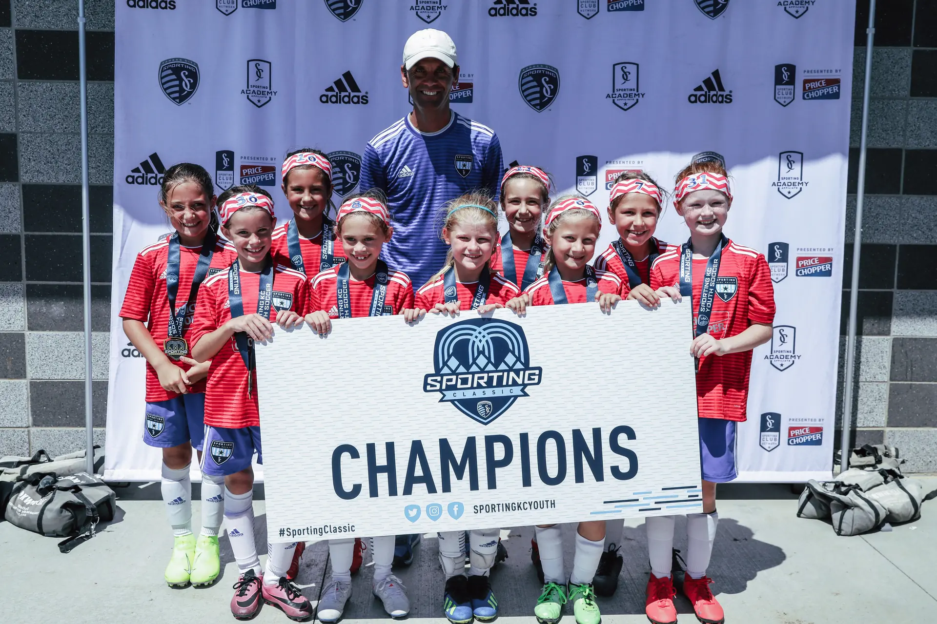 skcn0585-1920w-75121c08 A girls youth soccer team in red uniforms smiles with their coach, holding a Sporting Classic Champions banner after winning a tournament.