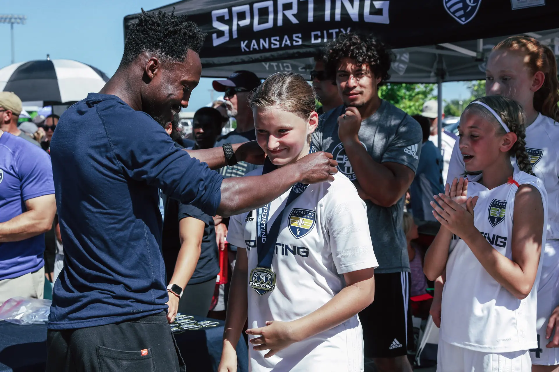 skcn0870-1920w-16ff0873 Sporting Kansas City staff member places a medal around a youth player’s neck as teammates applaud during an outdoor soccer awards ceremony.