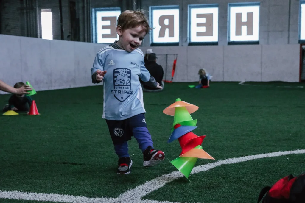 A young boy wearing a light blue "Sporting Stripes" soccer jersey and dark pants joyfully kicks a tall stack of colorful training cones on an indoor turf field. The cones are flying in different directions from the impact. The boy has a big smile and appears to be having fun. In the background, other children and adults are participating in activities, and large windows display the word 'HERE' in reverse due to the interior view.