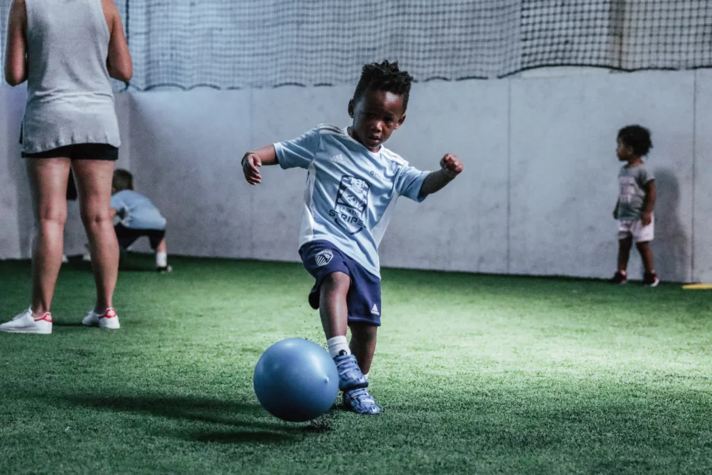 A young child wearing a light blue soccer jersey and navy blue shorts is playing with a blue ball on an indoor turf field. The child is in mid-motion, appearing to kick the ball with focus and determination. In the background, other small children and an adult are also present on the field. The walls are white with a netted upper section, and the lighting highlights the central child’s activity.