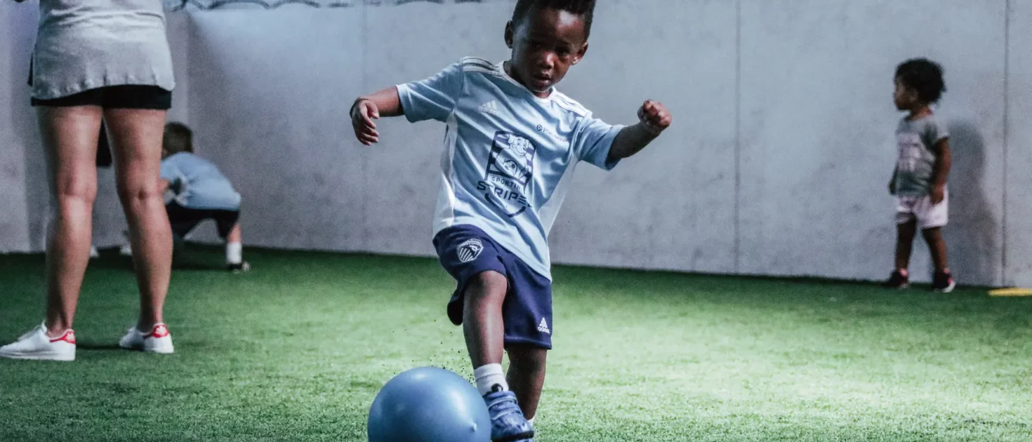A young child wearing a light blue soccer jersey and navy blue shorts is playing with a blue ball on an indoor turf field. The child is in mid-motion, appearing to kick the ball with focus and determination. In the background, other small children and an adult are also present on the field. The walls are white with a netted upper section, and the lighting highlights the central child’s activity.